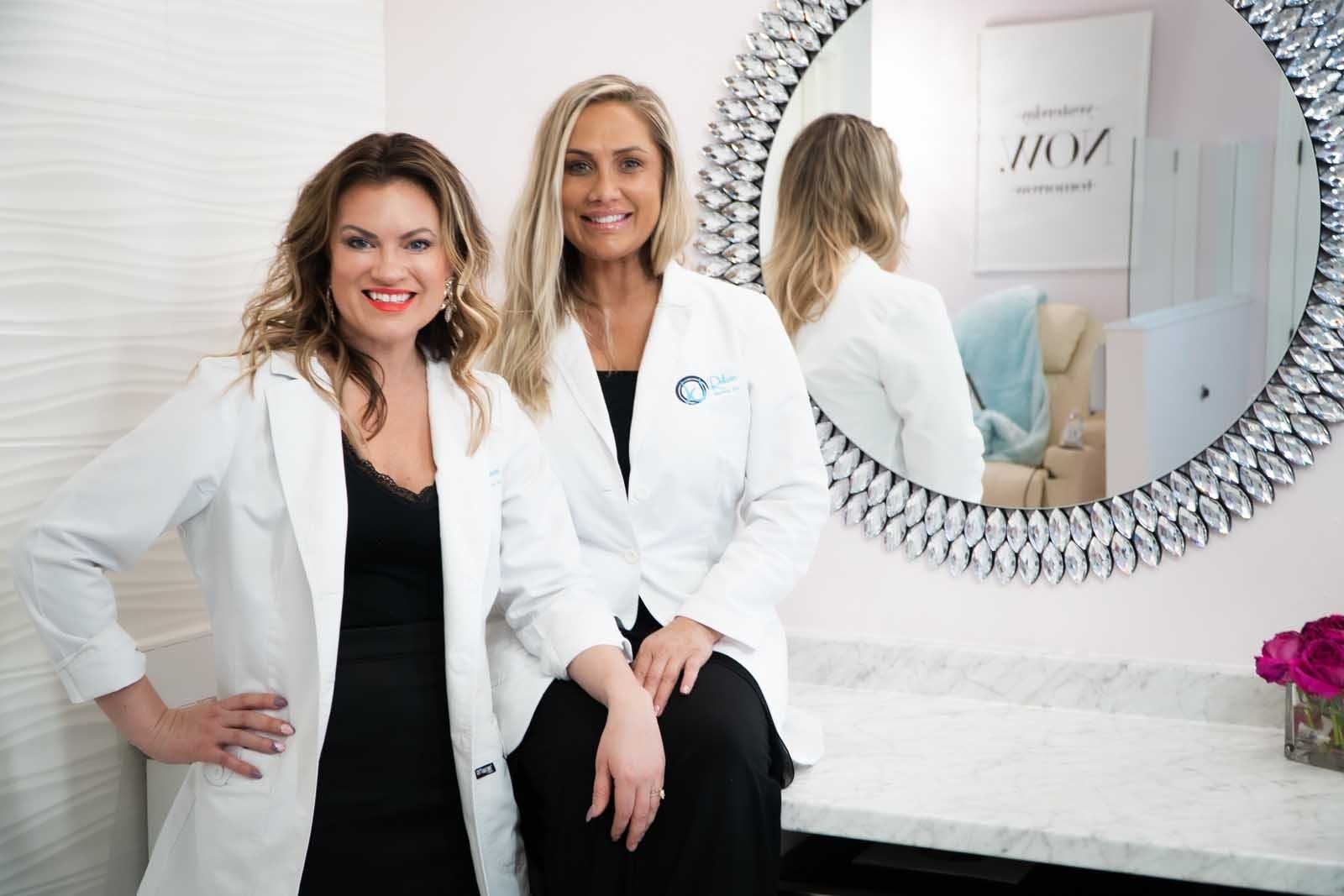Two women in white lab coats, smiling, near a mirror. Interior setting with pink accents.