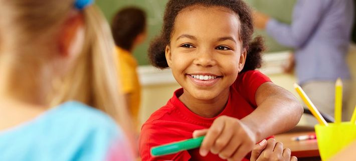 A young girl is sitting at a desk in a classroom holding a green marker.