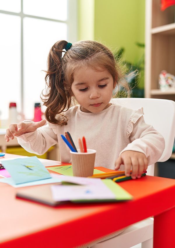 A little girl is sitting at a table playing with markers and paper.