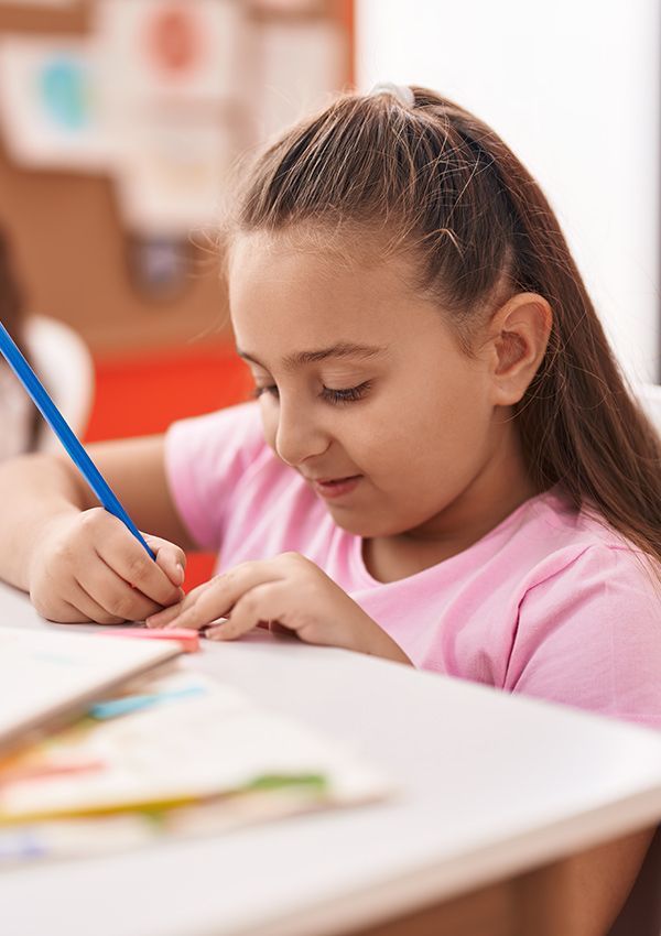 A little girl is sitting at a table drawing with a blue pencil.