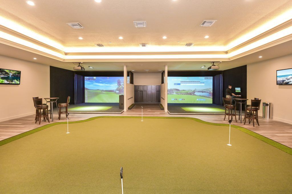 Indoor golf simulator room with two hitting bays, putting green, and screens displaying courses.
