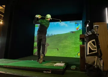 Man swinging golf club in an indoor simulator; green screen displays a golf course.