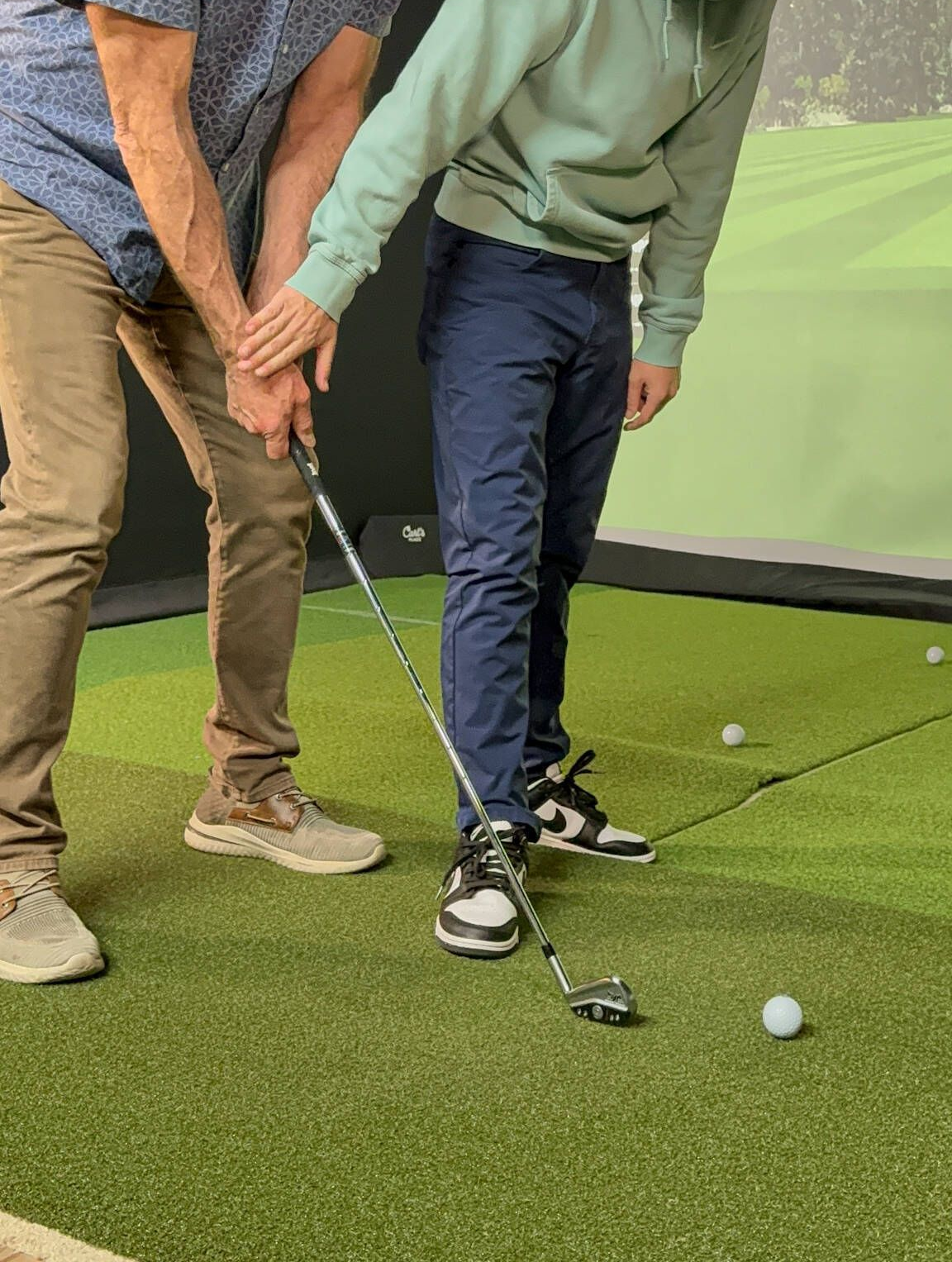 Two people practicing golf in an indoor simulator. One person assisting the other with the club grip.