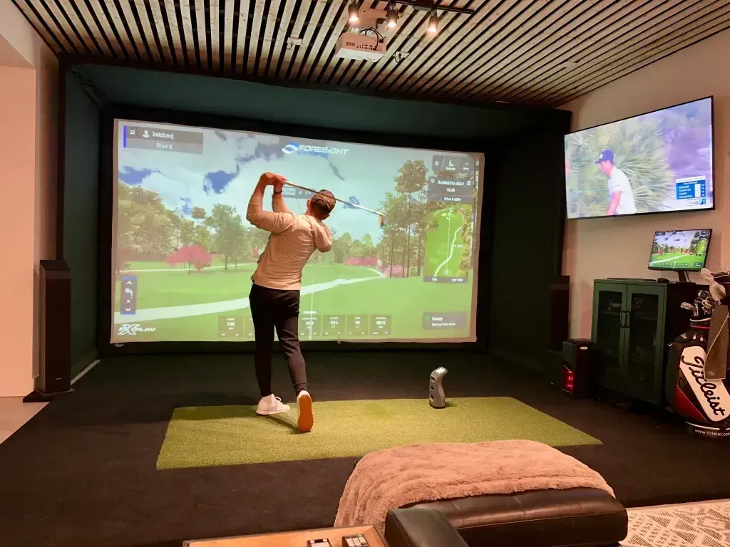 Man swinging a golf club in a home golf simulator, striking a ball. Green screen shows a golf course.