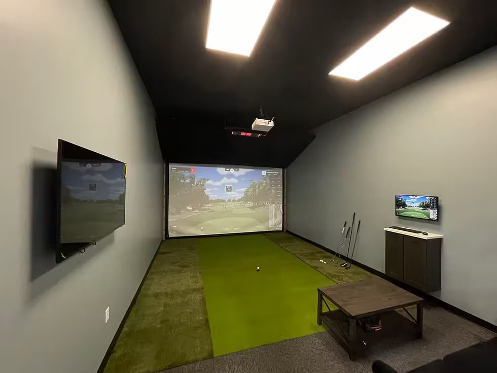 People playing golf: a woman swings, two others watch. Green field, trees in the background. All wear black.
