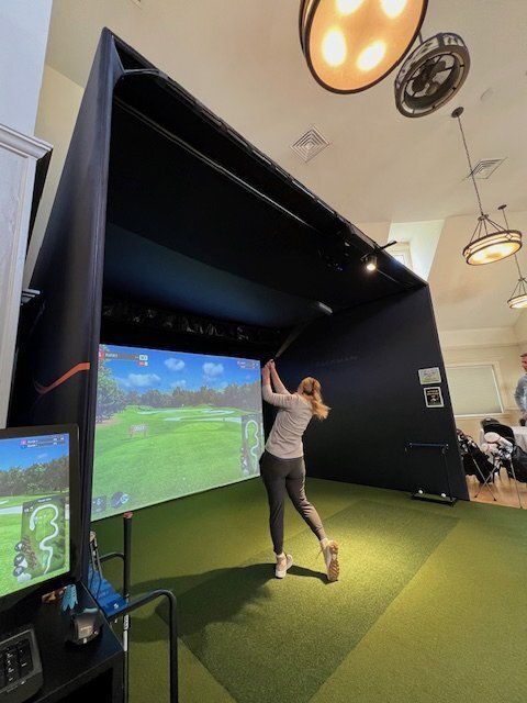 Woman swings golf club inside a golf simulator. Dark enclosure, green turf, screen displays golf course, overhead lighting.
