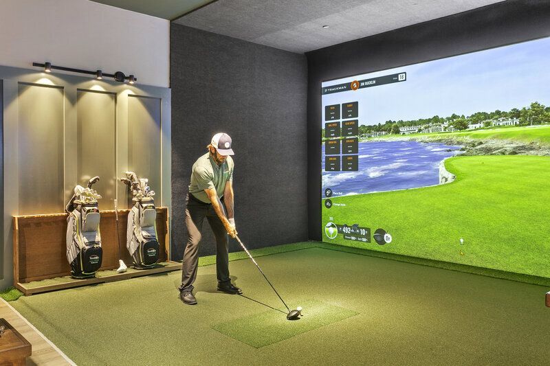 Man in golf simulator, swinging club toward a screen displaying a golf course. Interior setting, golf bags to the left.