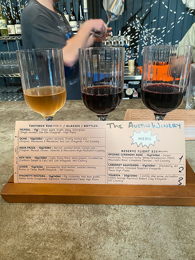Wine flight of three glasses — one white and two reds — at Austin Winery, with a branded menu in the foreground