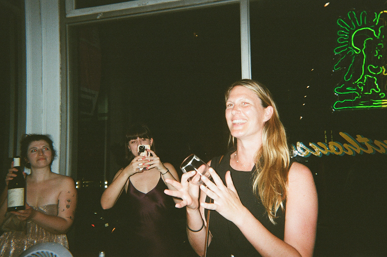 Woman speaking with two women behind her, one holding a wine bottle