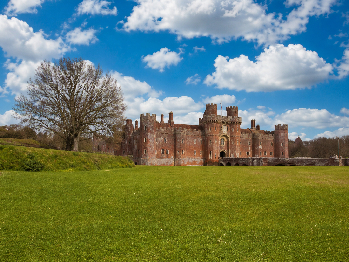 Red-brick castle on a grassy hill under a bright blue sky with scattered clouds