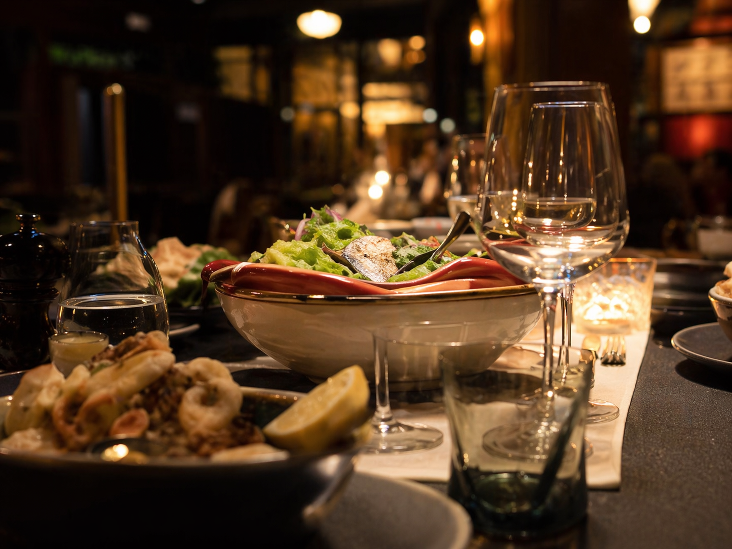 Dimly lit restaurant table with plated food, wine glasses, candles, and a salad bowl
