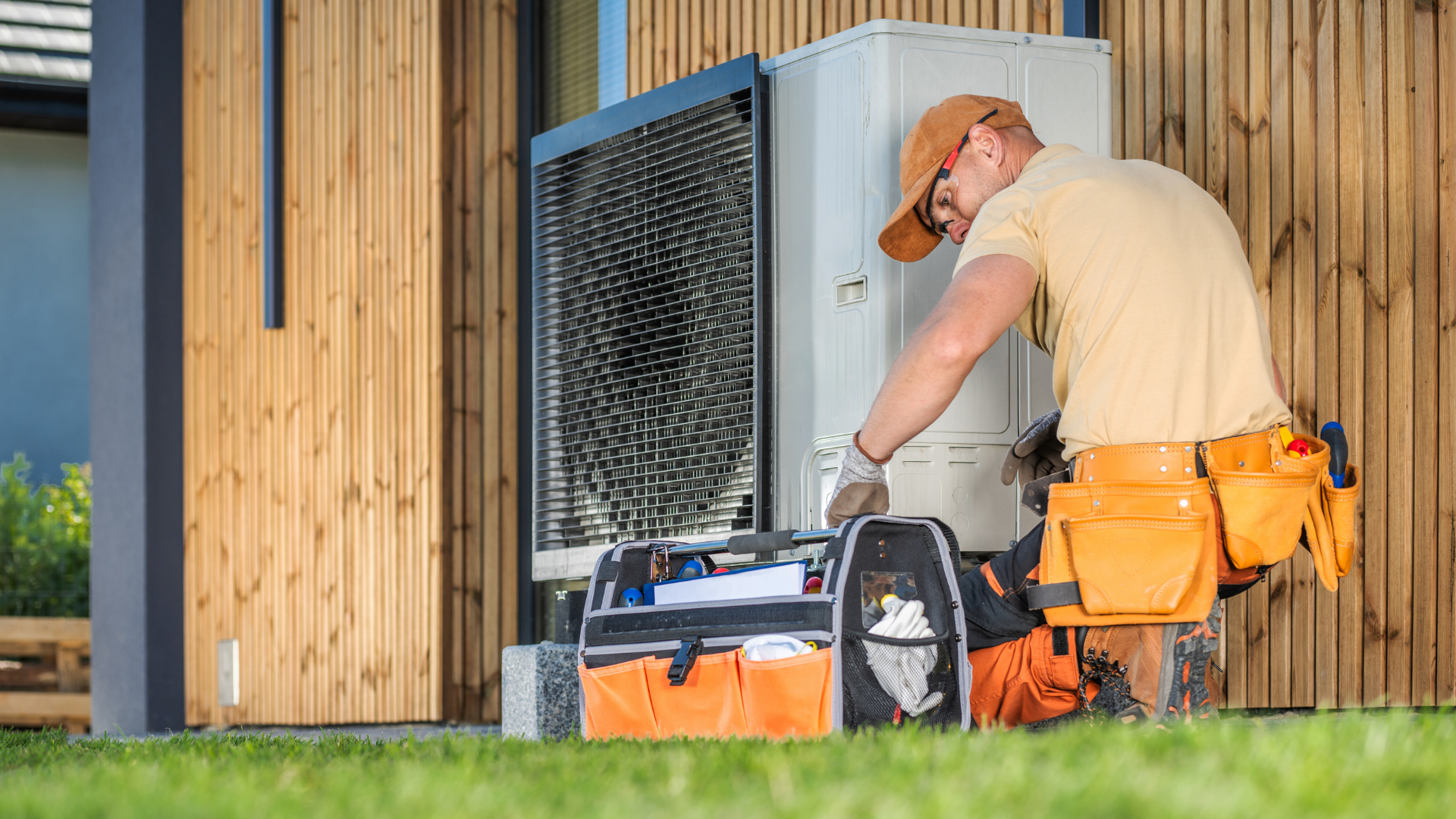 A technician working on an outdoor HVAC unit, wearing a tool belt, near a wooden building.