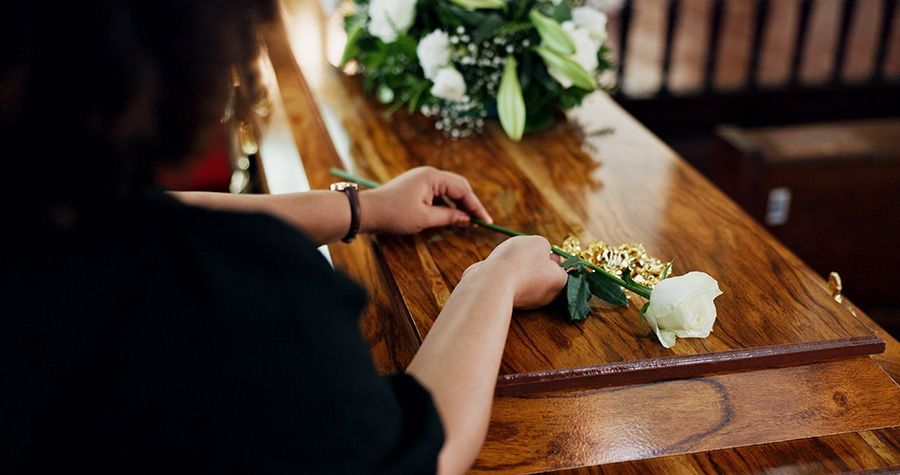 Person placing a white rose on a wooden casket with floral arrangement, indoors.