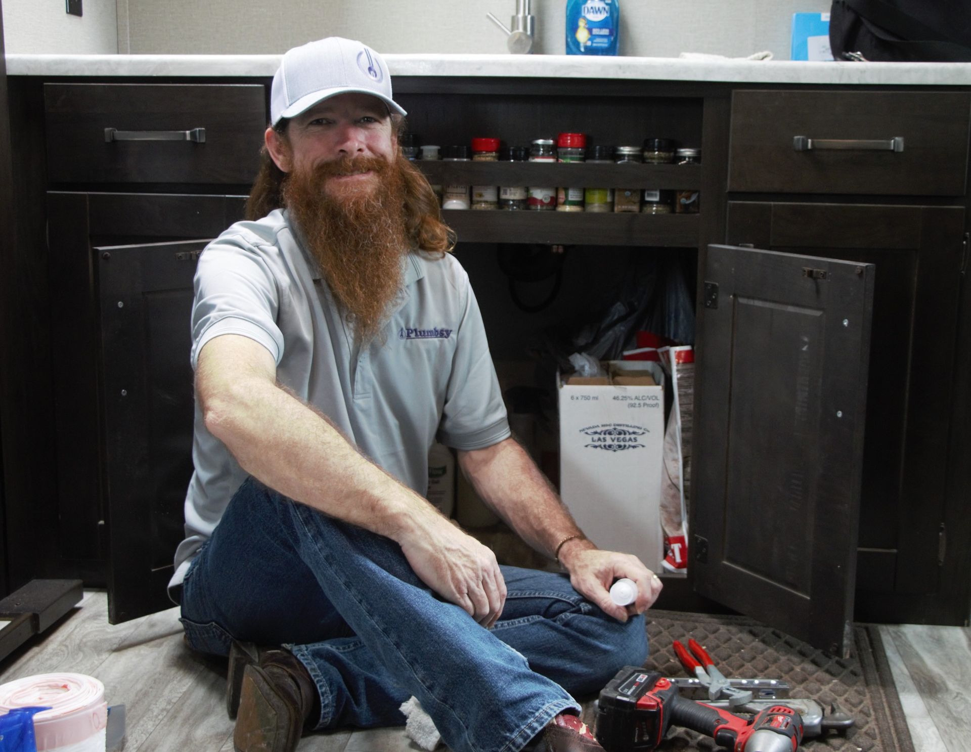 A man with a beard is sitting on the floor under a sink.