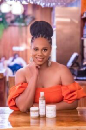 A woman in an orange top is sitting at a table with bottles of cosmetics.