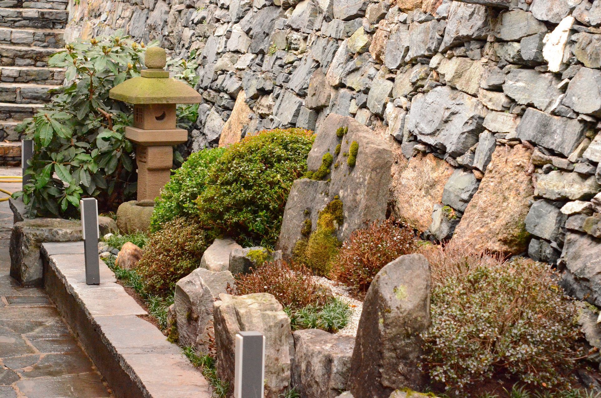 Un muro di pietra con una lanterna giapponese al centro, circondato da piante e rocce.