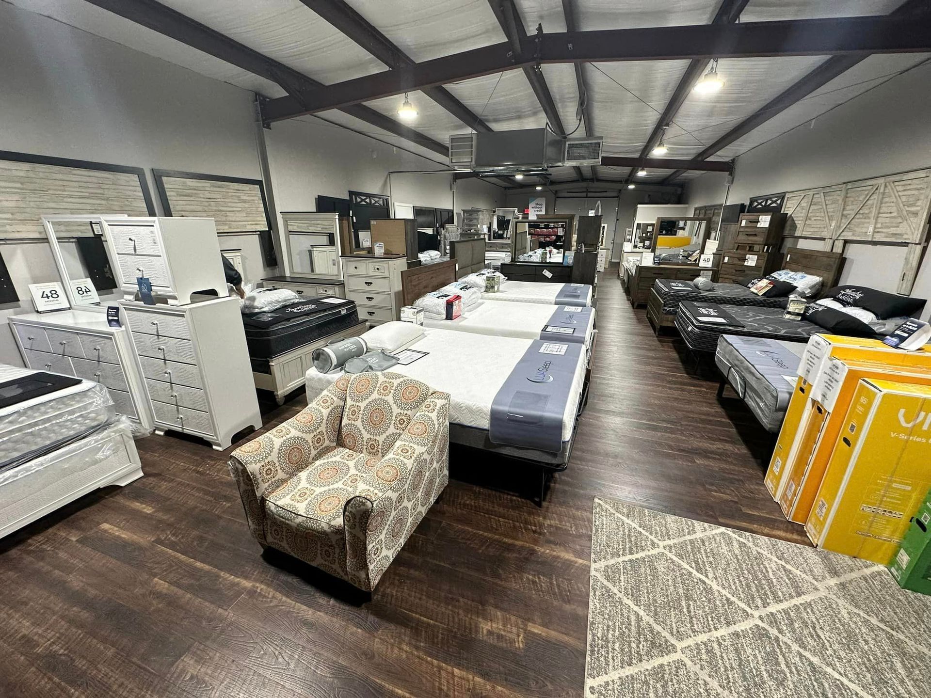 Furniture store interior with multiple beds, dressers, and a patterned armchair on a wood floor.