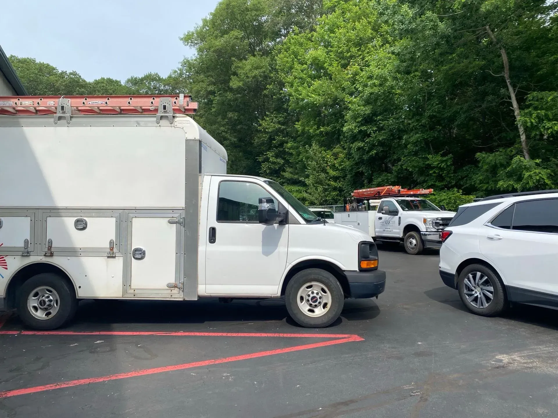 White utility truck parked in a lot beside a white SUV, with trees and other work vehicles in the background