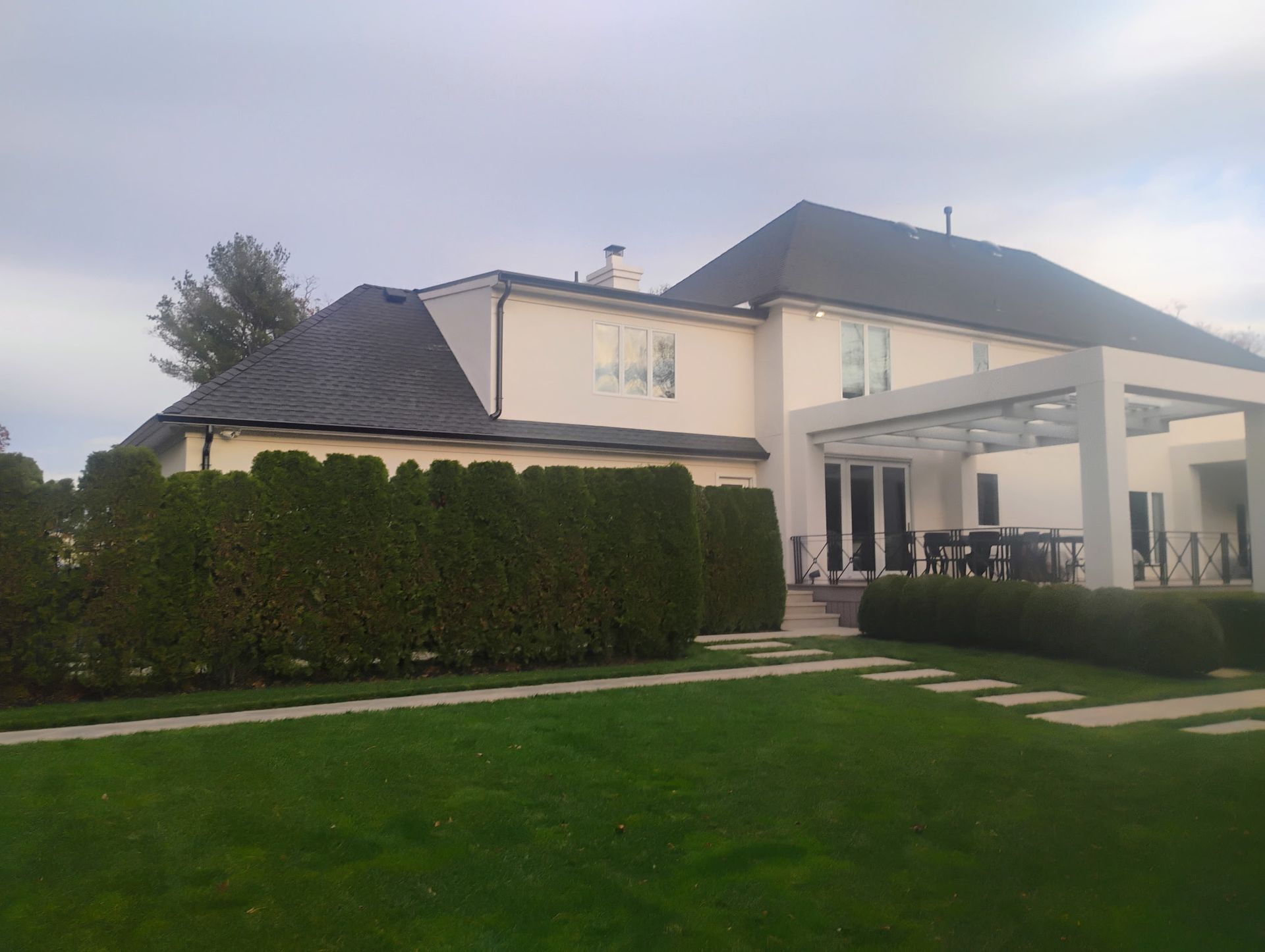 Two-story house with black roof, white walls, and a patio, surrounded by green grass and hedges, under a cloudy sky.