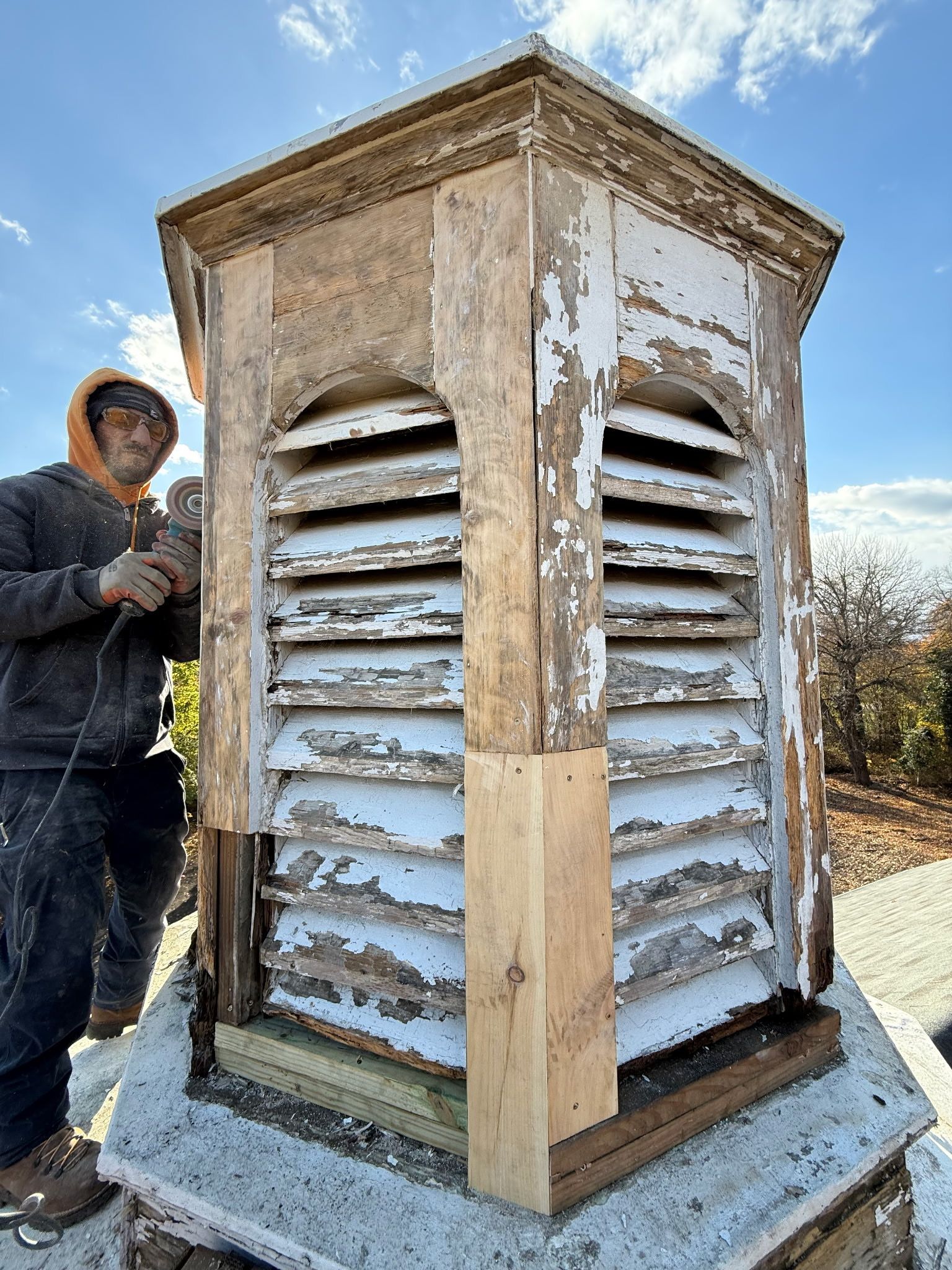 A person works on a weathered, white-painted louvered cupola on a rooftop. Bright sky in background.