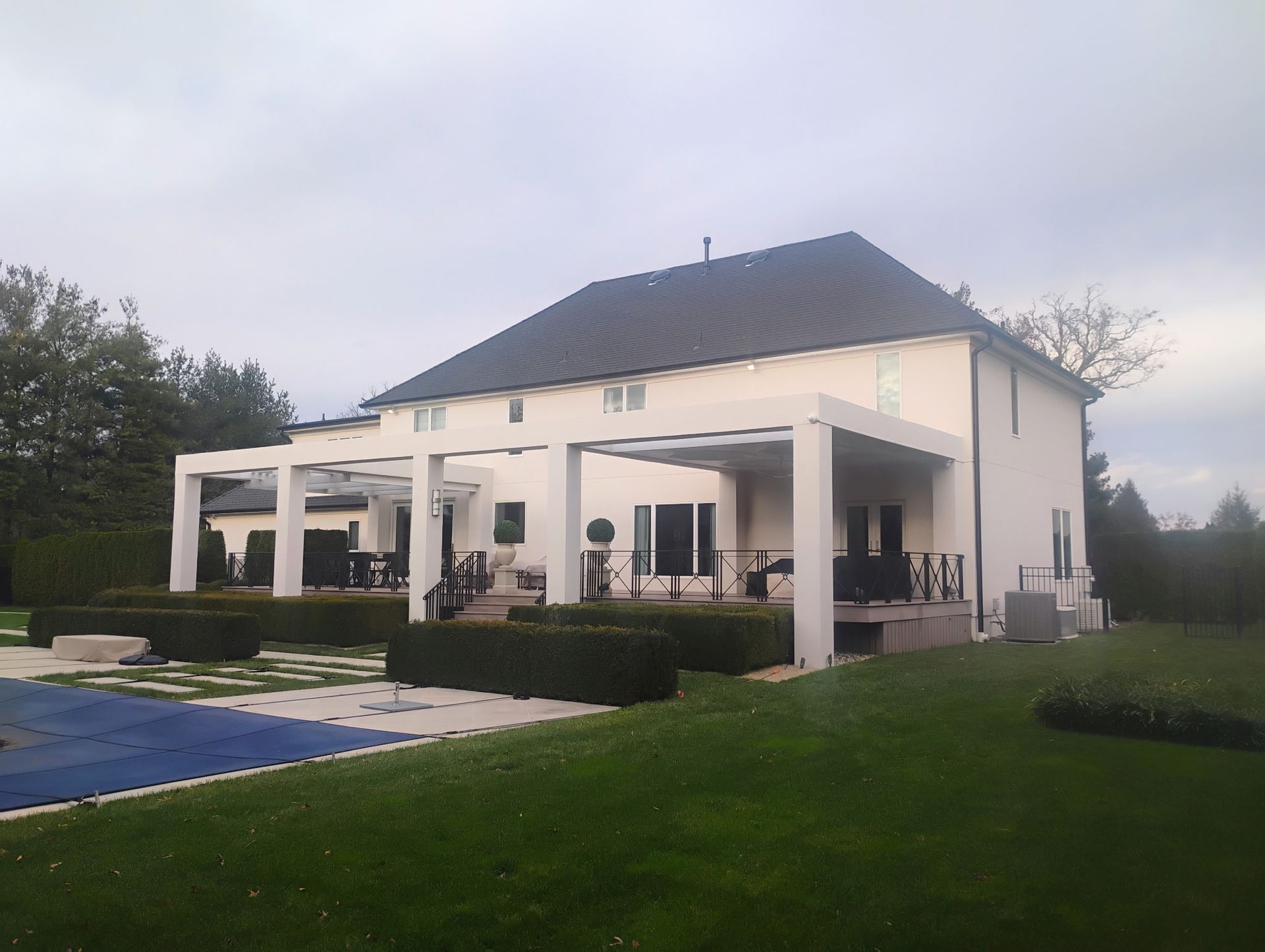 Beige two-story house with white pergolas, a pool, and green lawn under an overcast sky.