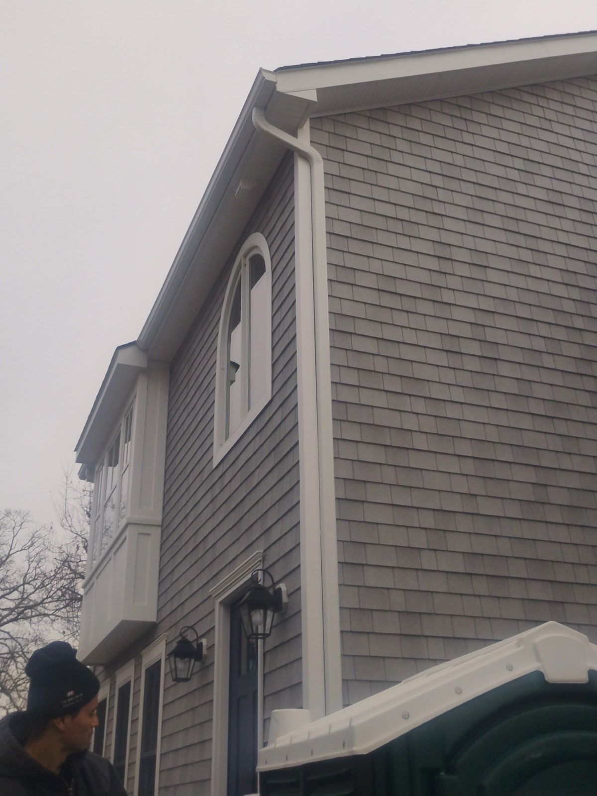 Exterior of a two-story building with gray shingles, white trim, and a curved window. A person stands nearby.