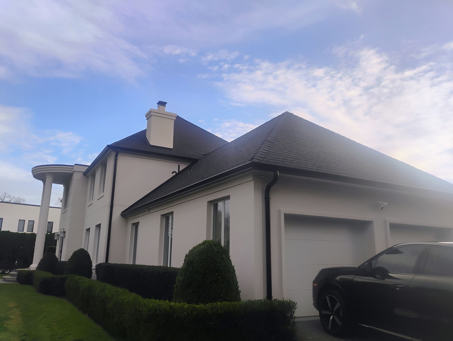 White two-story house with black roof, columns, and garage. Black car parked in front. Blue sky.