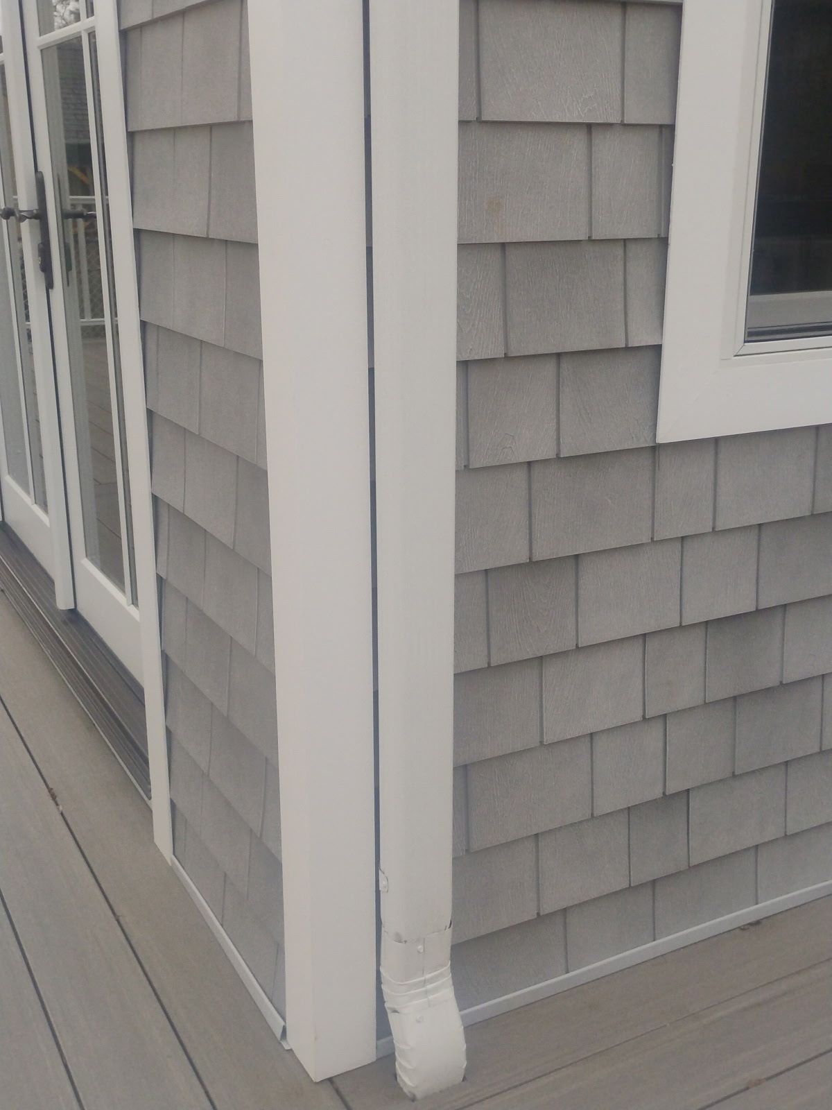 Gray shingle siding with white trim and a downspout, next to a deck and window frame.