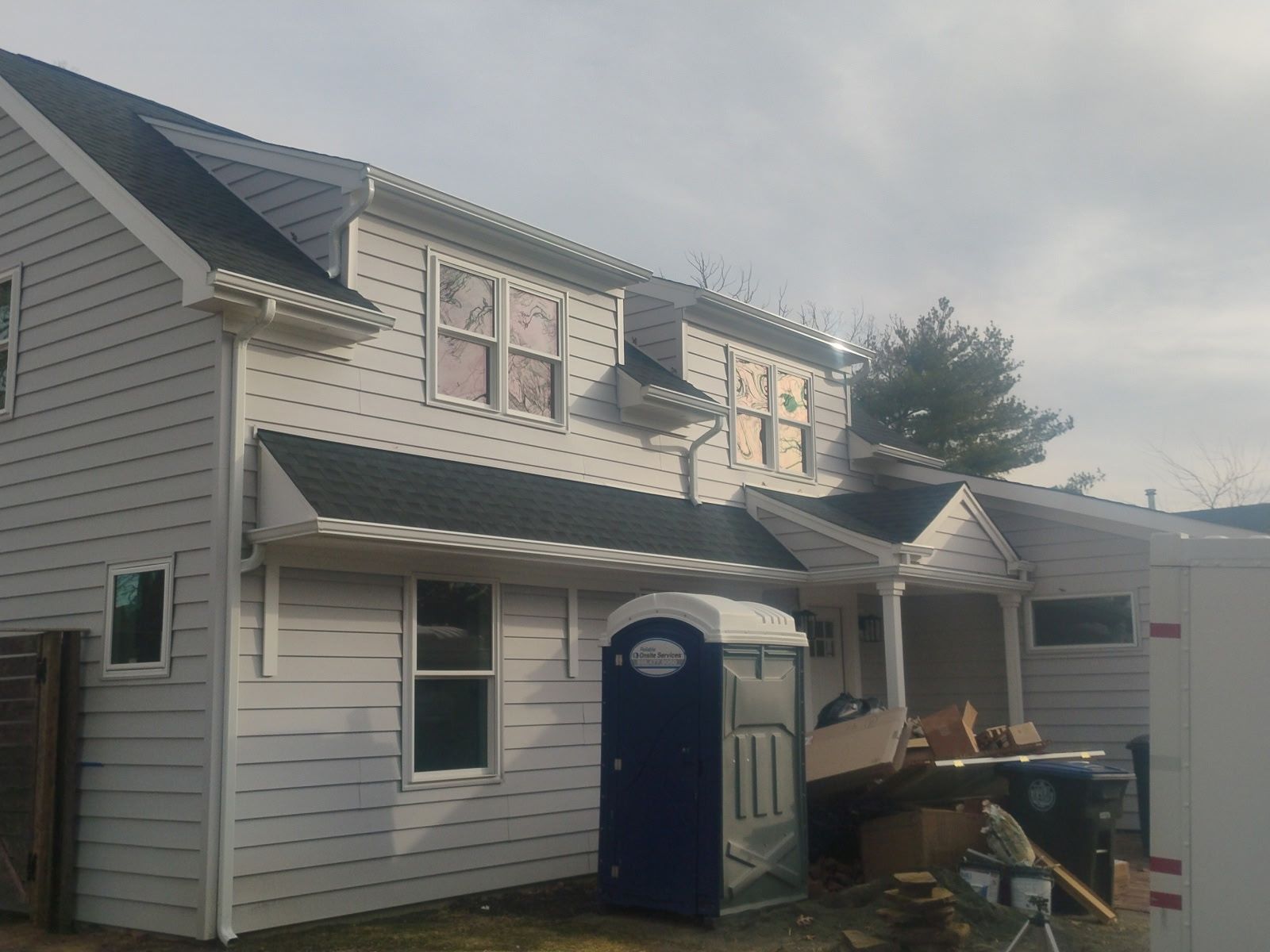 Two-story house with gray siding, black roof, and a blue portable toilet in front. Construction debris visible.