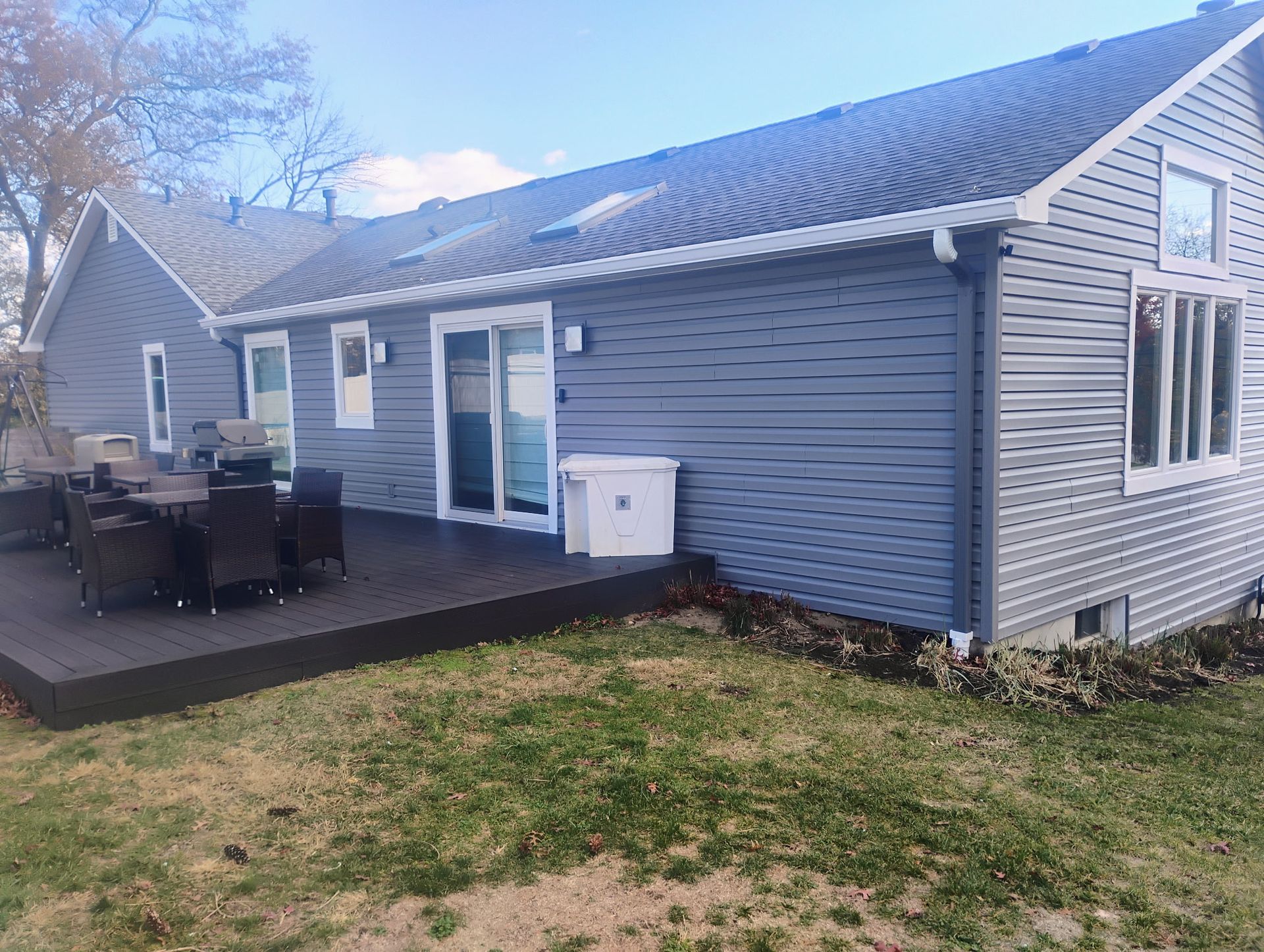 Gray house with deck, outdoor furniture, and sliding glass door, on a grassy lawn under a blue sky.