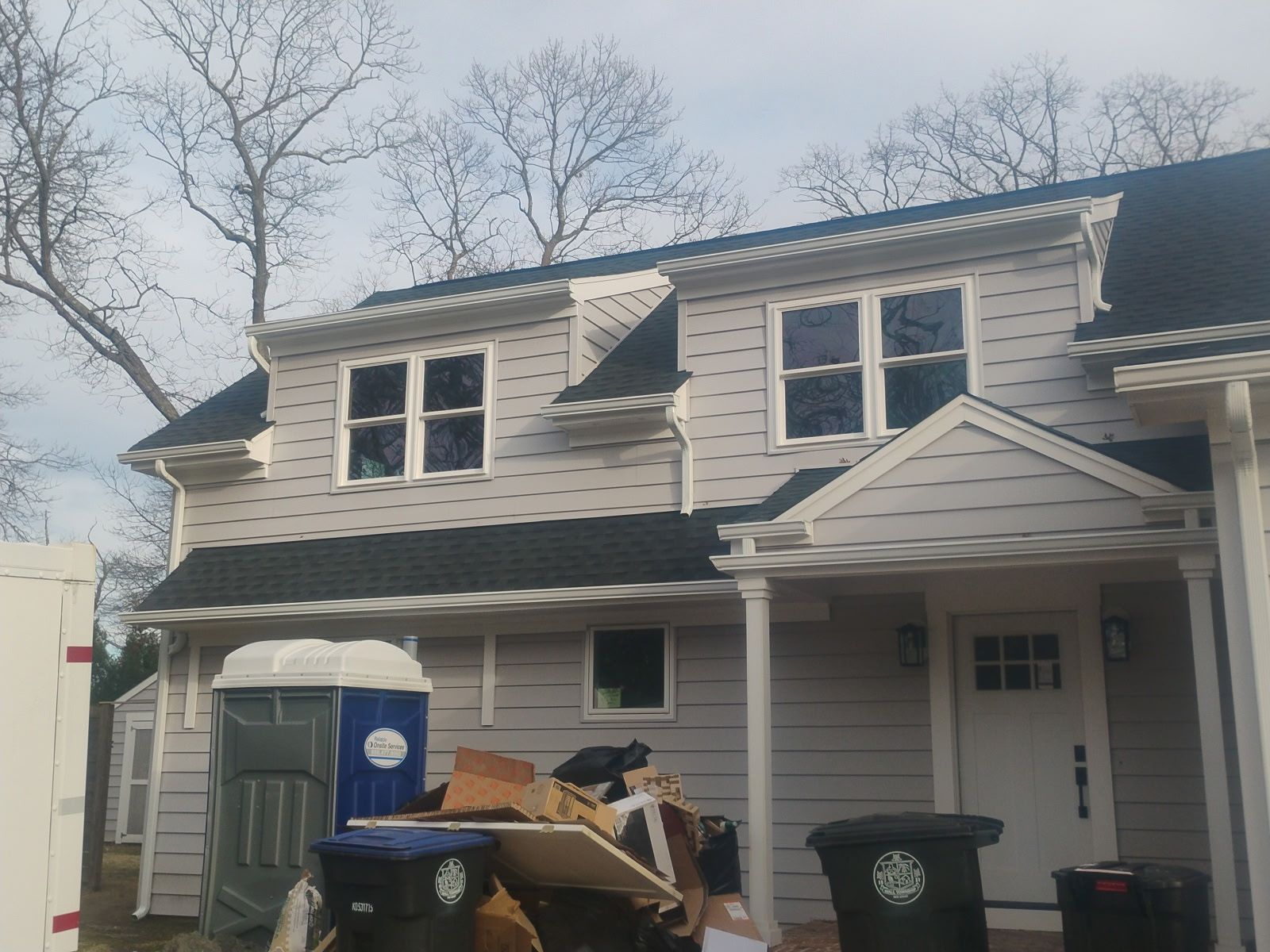 Two-story gray house with black roof, small port-cochere, and waste bins outside.