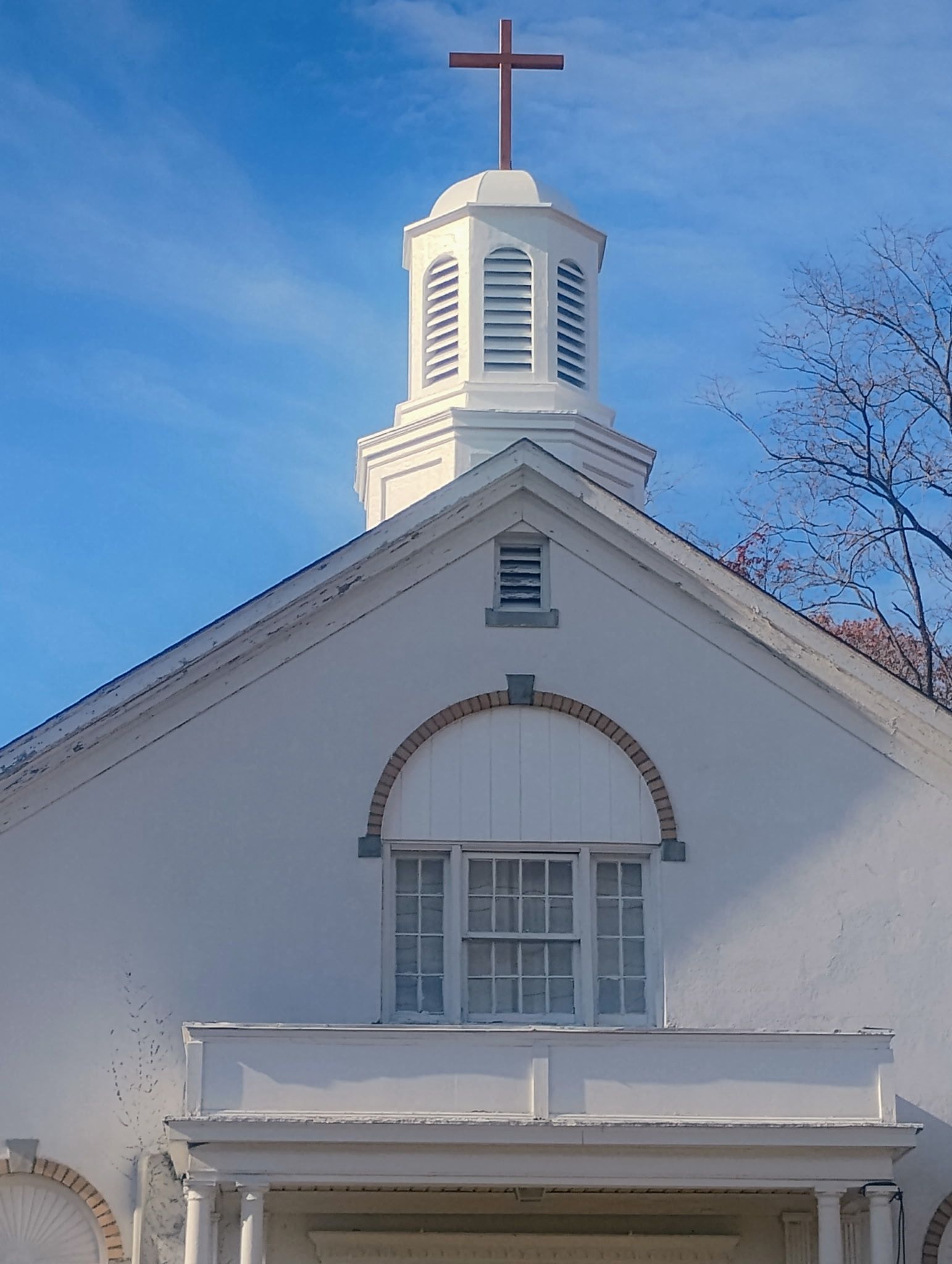 White church building with steeple, cross, and arched window against a blue sky.