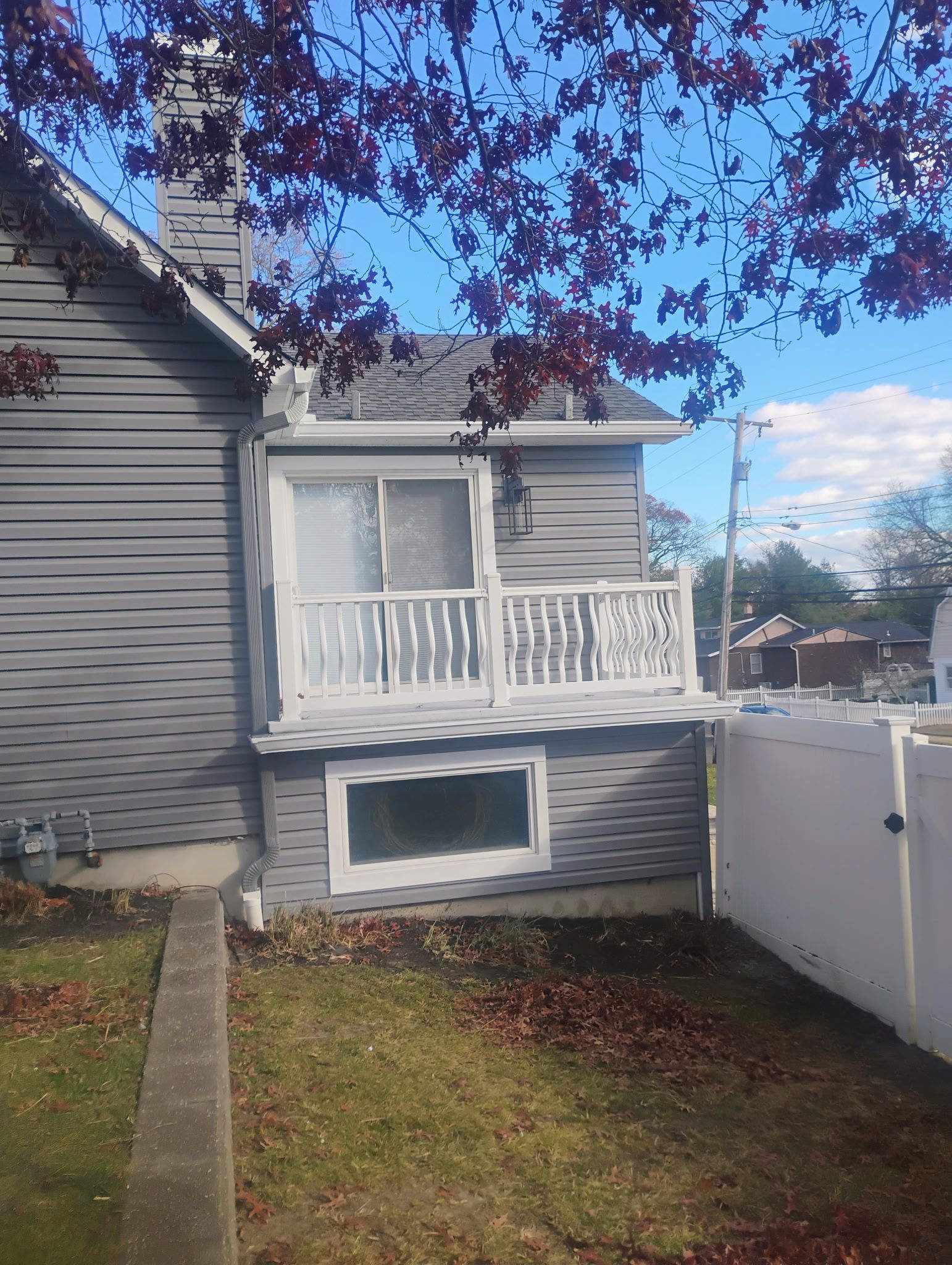 Gray house with white balcony, chimney, window, and small yard with fall leaves.