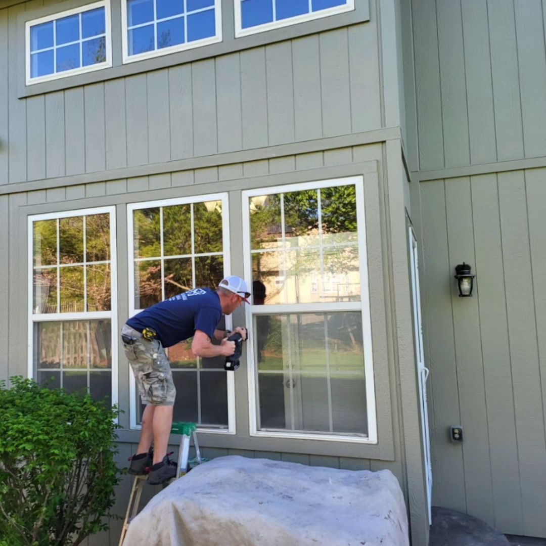 A man is standing on a ladder working on a window on the side of a house.