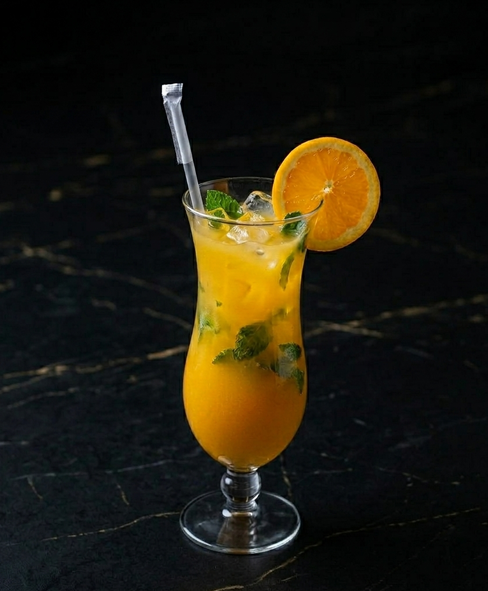 A tall glass of orange cocktail with ice, mint leaves, and an orange slice garnish, set against a dark marble background.