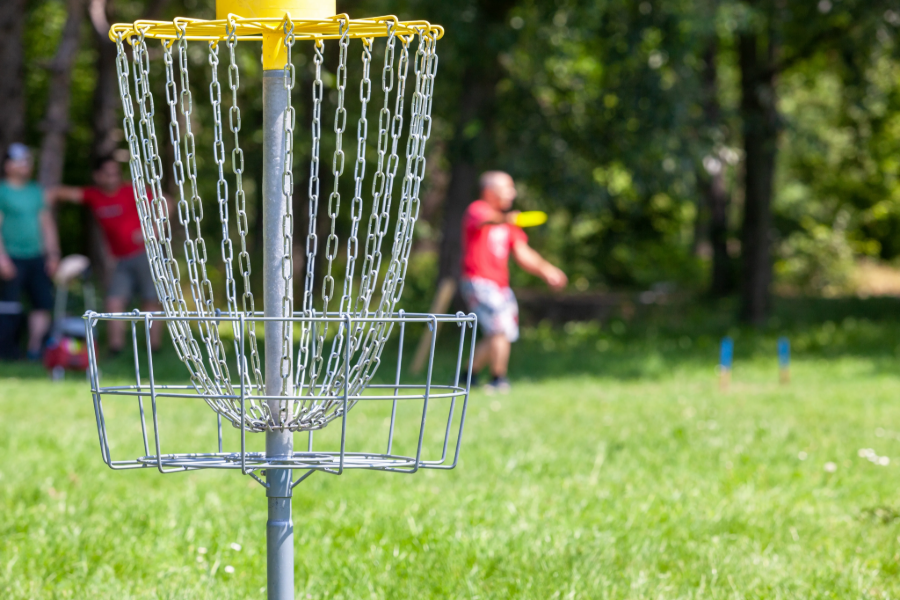Disc golfer mid-throw toward a basket on a grassy course, with sponsor banners behind him