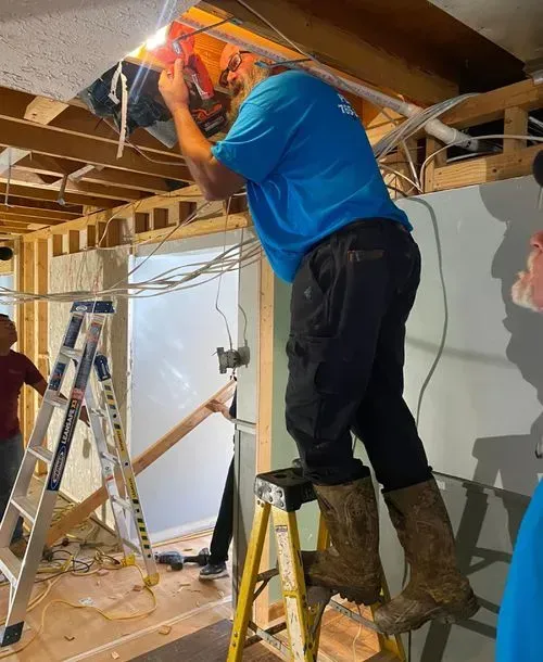 Man in blue shirt, boots, using a saw on ceiling electrical wiring while standing on a step ladder in a room under construction.