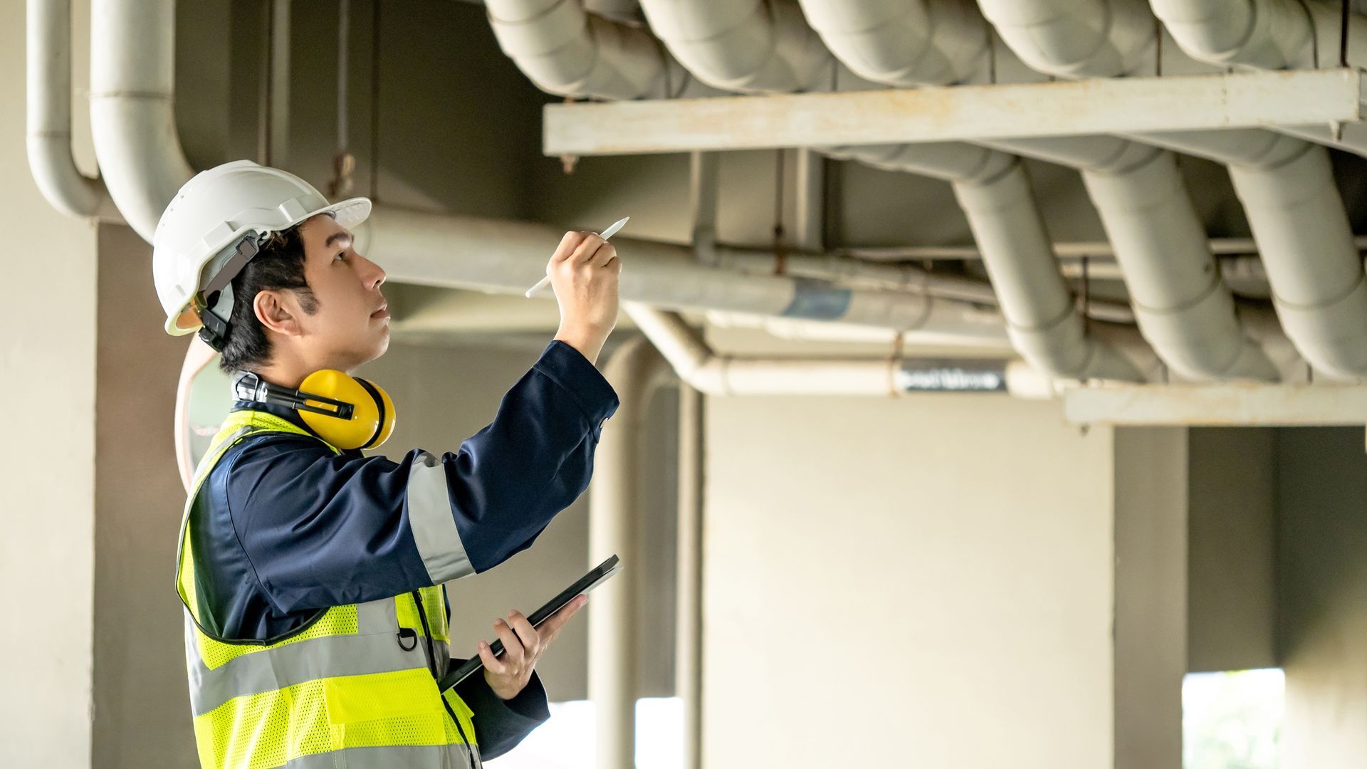 Construction worker in hard hat and safety vest inspecting overhead pipes, holding tablet and pen.