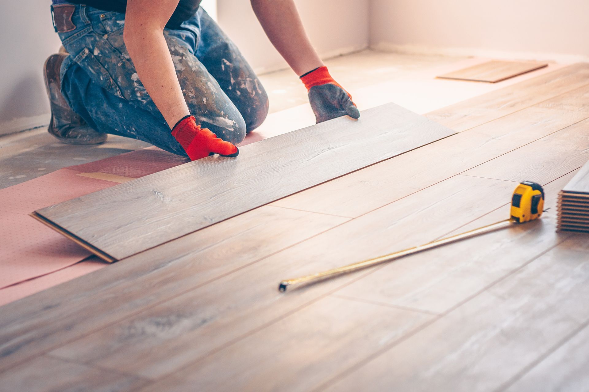 Person installing wooden floorboards, wearing gloves, kneeling in a room.