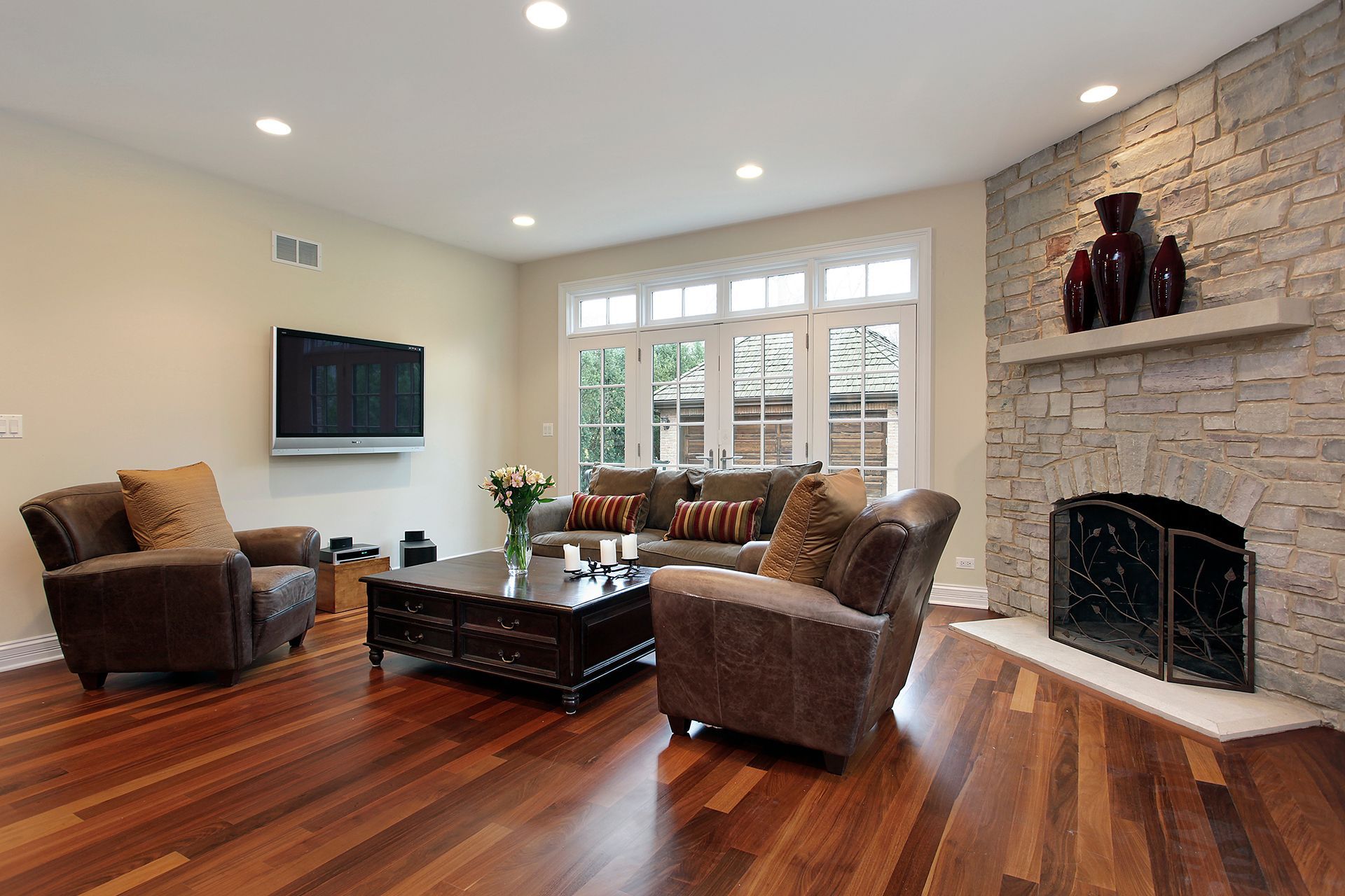 Living room with wood floors, stone fireplace, brown furniture, and large windows.