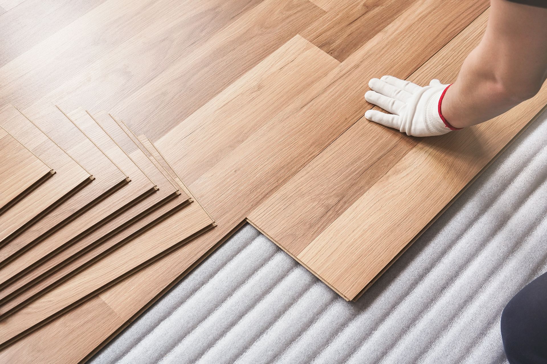 Person wearing gloves installing wooden floorboards on white underlayment.