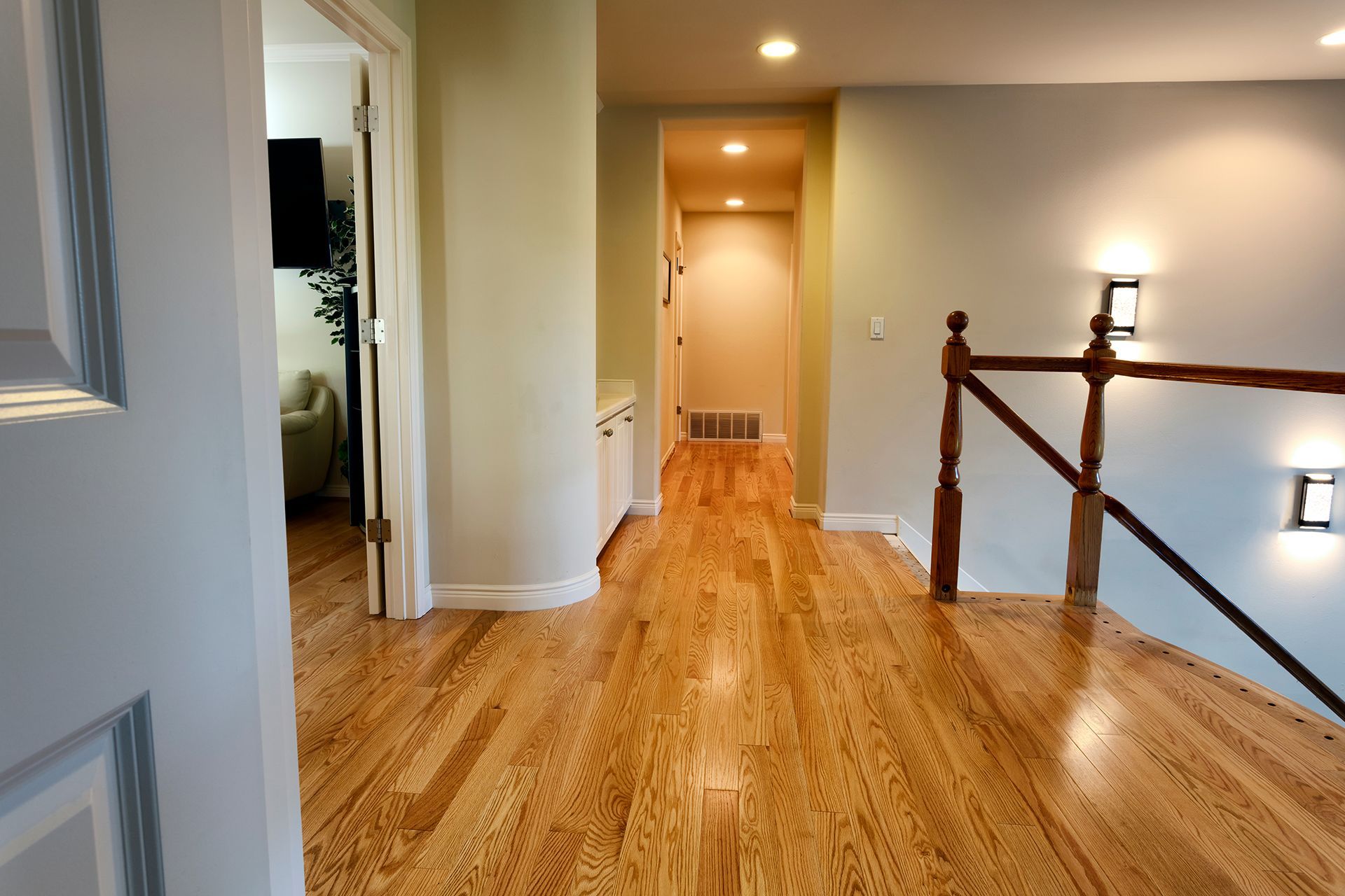 Hallway with hardwood floor, neutral walls, and a staircase railing. Doorway to a living room on the left.