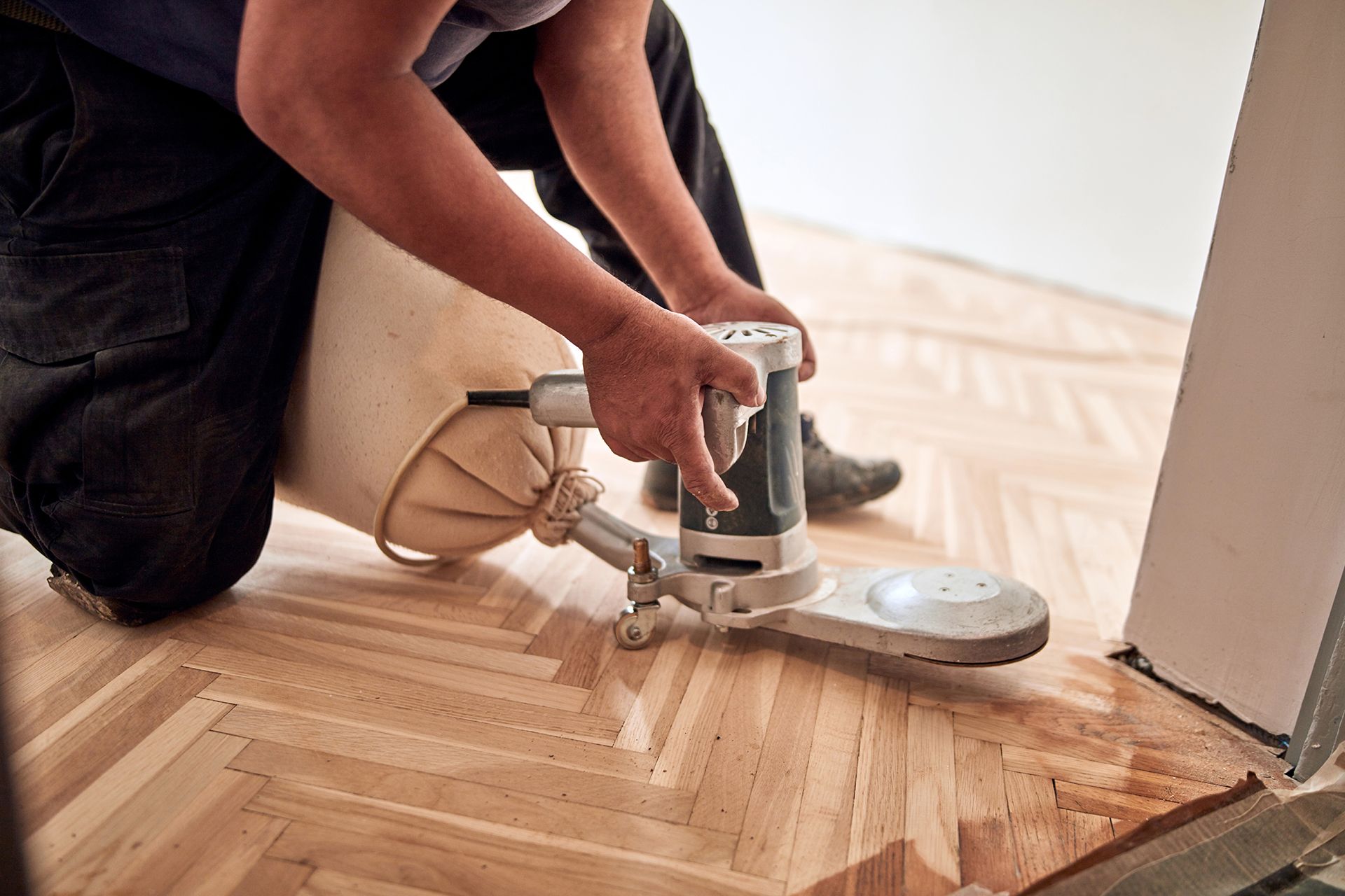 Person sanding parquet flooring with a disc sander, dust bag attached.