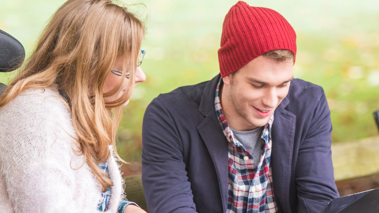 A man and a woman are sitting on a bench looking at a laptop.