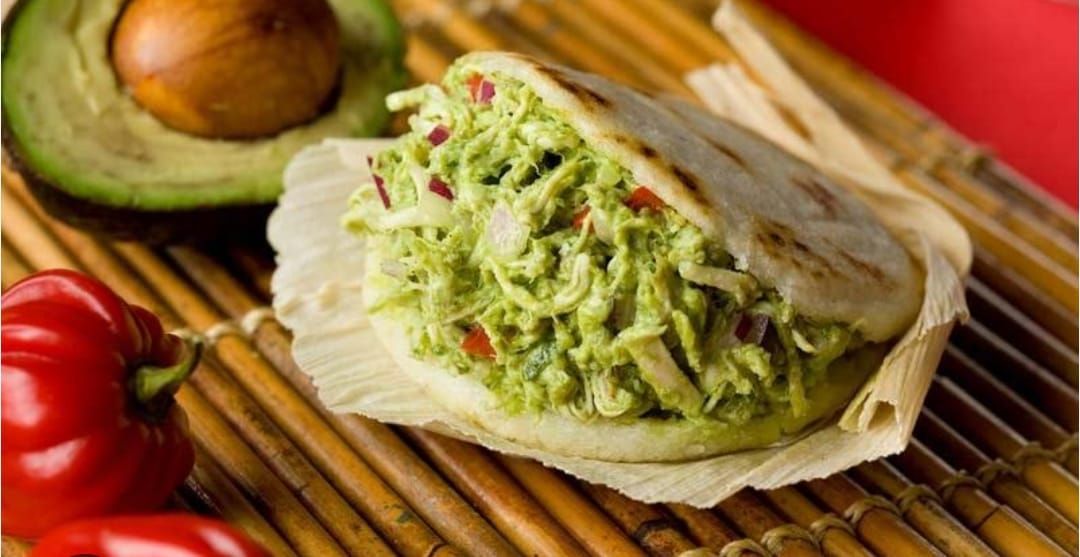 Arepa rellena de aguacate verde y ensalada de pollo, con medio aguacate y un pimiento rojo al lado.