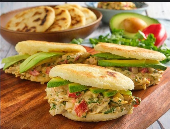 Sándwiches de arepas con aguacate y relleno sobre una tabla de madera, con más arepas y aguacate visibles en el fondo.