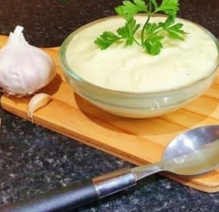 Alioli de ajo en un recipiente de vidrio con guarnición de perejil, cabeza de ajo y cuchara sobre una tabla de madera.