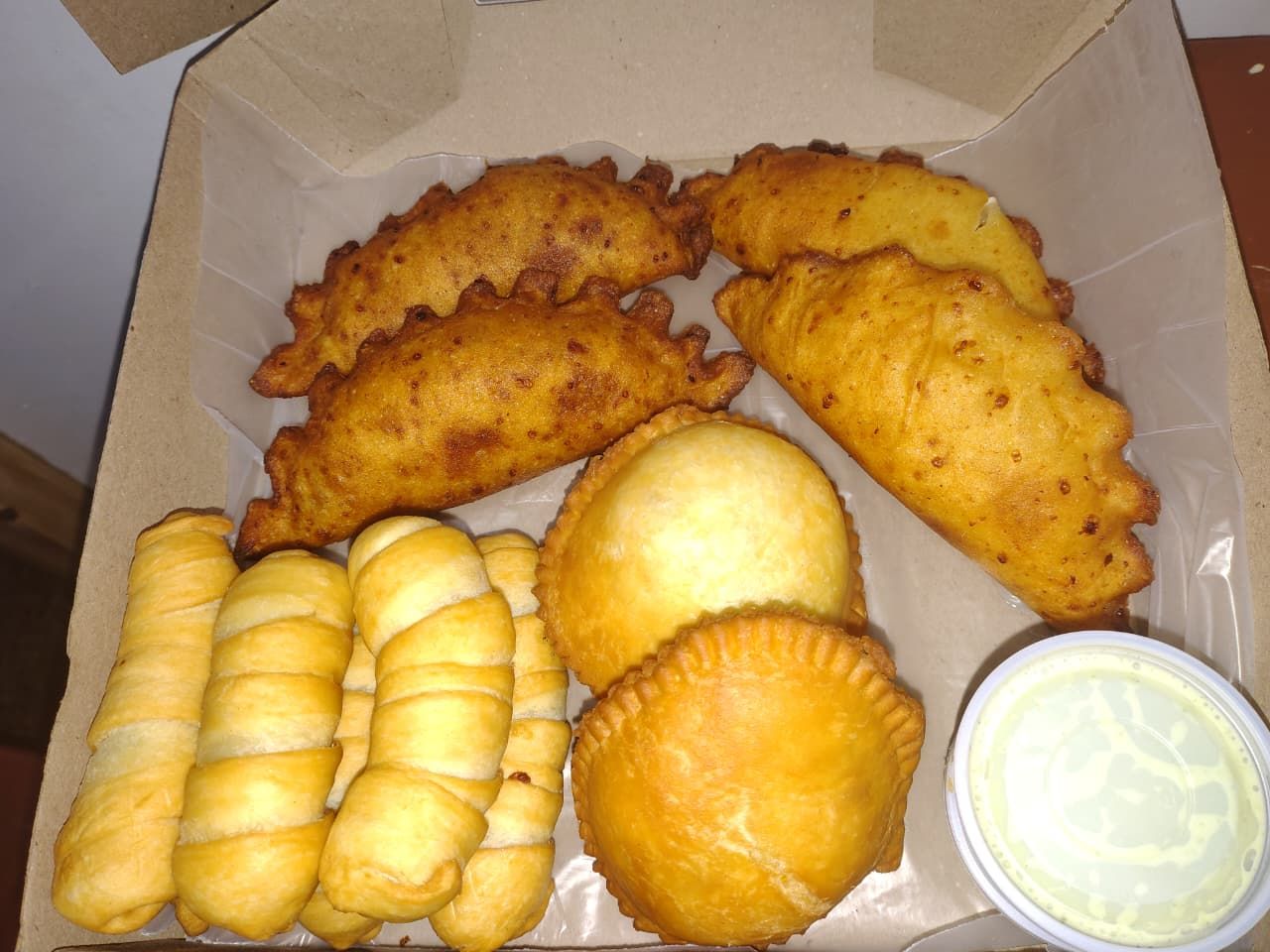 Una caja de pasteles fritos dorados y tequeños con un poco de salsa para mojar.