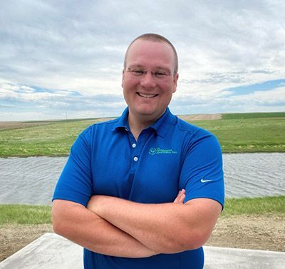 A man in a blue shirt is standing with his arms crossed in front of a body of water.