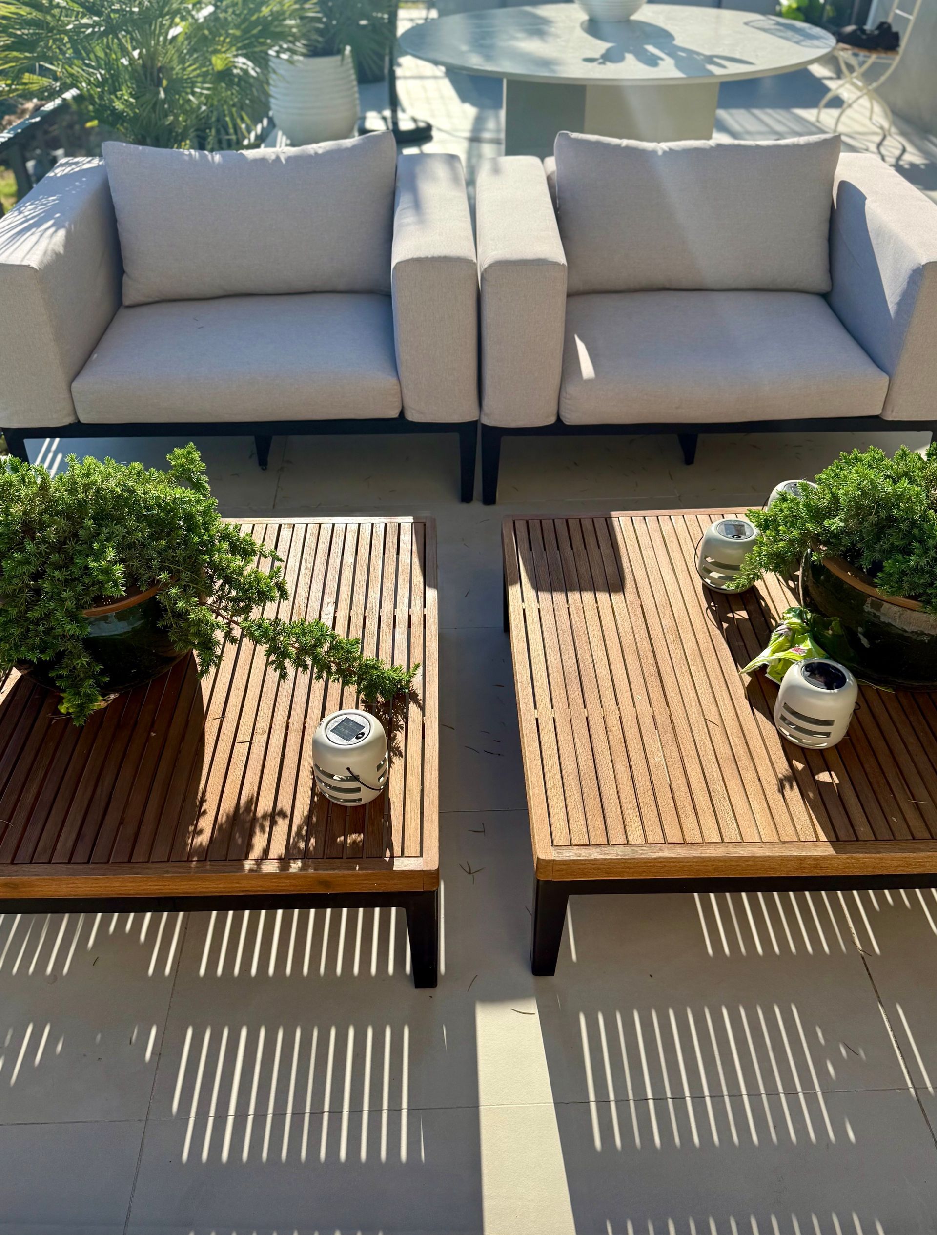 Two outdoor chairs with light cushions face two wooden tables with plants, sunny setting.